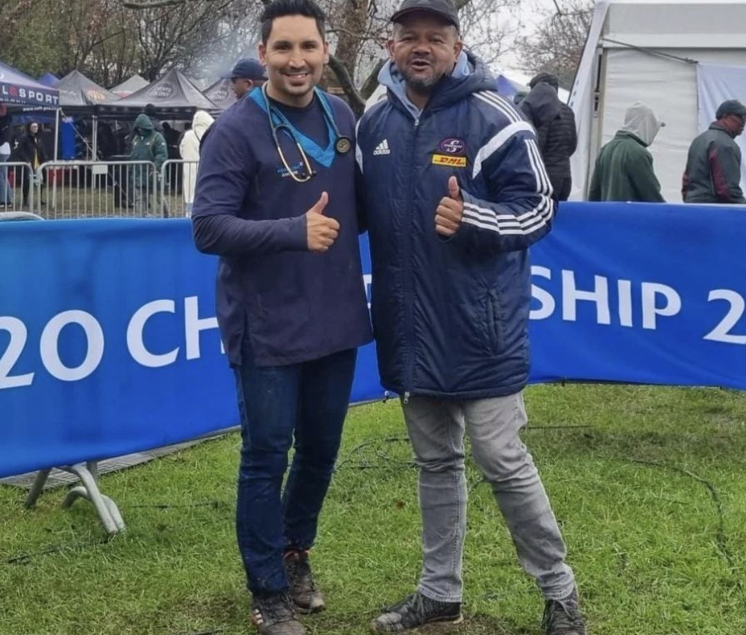 Dr Arthur Williams on the right in a navy padded jacket with a colleague giving thumbs up beside a rugby championship banner